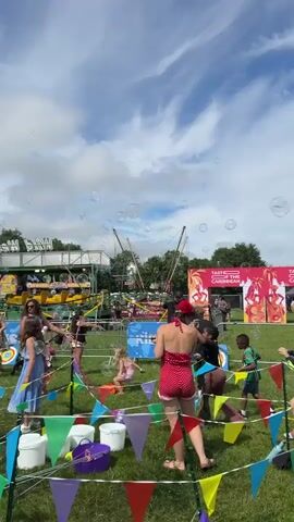 This is a vertical, outdoor video taken on a sunny day with some clouds. The scene appears to be a fair or carnival. In the foreground, several children and adults are visible. Some children are holding bubble wands and blowing bubbles, which are floating in the air. There are colorful pennant banners strung up in the grass. In the middle ground, there are various carnival rides, including a yellow ride with multiple cars and a bungee trampoline. Fencing separates the public area from the rides. In the background, trees and a blue sky with white clouds are visible. The overall atmosphere seems festive and lively, with people enjoying outdoor activities.
