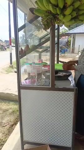 A person is standing behind a food cart and chopping ingredients with a knife. The food cart has a glass display case with various items inside, including bottles and containers. A bunch of green bananas hangs from the top of the cart. The cart is situated outdoors next to a road, with buildings and trees visible in the background. The ground appears to be dirt and grass.