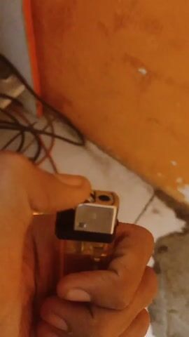 A tan hand holds a transparent, light orange lighter with a silver top against an orange wall and speckled white and gray floor. The person ignites the lighter with their thumb, producing a small, bright yellow flame. They hold the flame steady for a few seconds before releasing the ignition and extinguishing it. Electrical wires are visible in the blurred background.