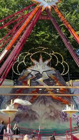 The video shows a large amusement park ride with a dragon theme. The ride is red and yellow and has lights. It is shaped like a pendulum with a dragon on the bottom. There are trees in the background. Several people are standing around the ride. The ride appears to be stationary.