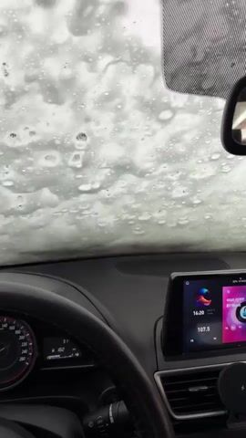 The video shows the interior of a car going through an automatic car wash. The view is from the driver's seat, looking through the windshield, which is covered in soap and water. Large brushes pass over the windshield, cleaning it. The dashboard and steering wheel of the car are visible, along with the car's infotainment system, which is displaying a colorful interface with a radio station playing. The car is stationary.
