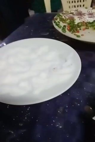 A close-up shot shows a table with several plates of food. One plate holds a white, cottony substance, another contains chopped green onions and red chilies in a clear liquid. The third plate has a large portion of mashed potatoes next to a mixture of what appears to be cabbage, meat, and gravy. A metal utensil is used to scoop some of the potato and other ingredients together. The table surface is dark and patterned. A white chair can be seen in the blurred background.