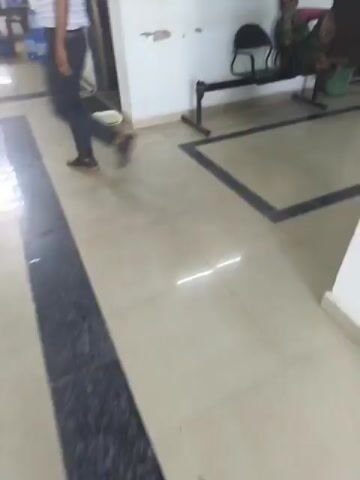 The video shows a long hallway with shiny tile floors that have a black stripe running down the center. The walls are white, and there are dark-colored doors on either side. Some windows with dark grilles are visible on the left side. At the beginning of the video, a woman is seen sitting on a bench. Further down the hallway, a red fire extinguisher is mounted on the wall near a chair. The video seems to be taken while walking down the hallway.