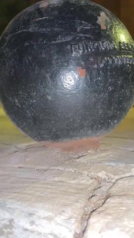 The video begins with a close-up shot of a black spherical object, possibly a cannonball, resting on a stone surface. Rust is visible on the object and the surface. The camera then pans upwards, revealing a building with a facade that appears to be a church or similar structure. The building is illuminated by warm-toned lights, creating a nighttime scene. The area in front of the building is paved with cobblestones. There are no people visible in the video.