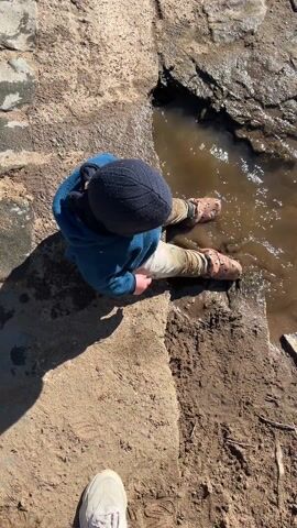 A young child, wearing a blue hooded jacket and dark blue beanie, is sitting and leaning towards a muddy puddle. The child is situated on a rocky, uneven ledge composed of light brown dirt, patches of darker soil, and embedded rocks. The child has both legs immersed in the murky water.  The water, reflecting sunlight, gives the impression of a shallow, stagnant pool bordering a rugged stone embankment. The child’s pants are multicolored, and their feet are clad in brown shoes. A pair of white sneakers is visible at the bottom of the frame, implying the presence of a person holding the camera. The surrounding environment appears to be an outdoor, possibly natural area.