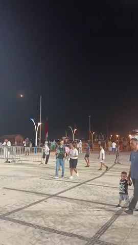 At night, a group of people are gathered in an outdoor area, likely a plaza or promenade. The sky is dark, with a faint moon visible. Several tall lampposts illuminate the scene, some with decorative lights. In the foreground, a young man wearing a t-shirt and jeans, holding a pink object, walks with a young woman in a striped shirt and skirt. Behind them, other people are milling about. There are metal barriers set up, and in the distance, a crowd is seated or standing. Flags and a building are also visible in the background. The ground is paved with large rectangular stones.