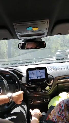 This is a first-person perspective video from inside a car. The driver's hands are visible on the steering wheel, and the passenger's hands are visible in the center console area. A car is in front of them on a winding road with trees and foliage on either side. The car's infotainment screen is visible, displaying what appears to be navigation or audio information. There are two heart-shaped stickers, one blue and one yellow, on the overhead console. The video is shaky, indicating movement.