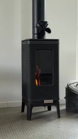 The video shows a black wood-burning stove with a glass door. Flames are visible inside the stove, and the word "INVICTA" is printed on the glass. A black pipe extends from the top of the stove, with a small, four-bladed fan attached to it. The fan is spinning. The stove is standing on a light-colored tiled floor against a white wall. A metal bucket sits to the right of the stove.