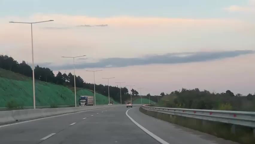 A forward-facing dashboard camera view shows a four-lane highway with two lanes in each direction. The camera is in the rightmost lane. The highway is bordered by green, grassy hills on both sides. Several streetlights are visible overhead. In the distance, a white car travels away from the camera in the same direction. On the left side of the road, two large trucks travel in the opposite direction. The sky is a light blue with some scattered clouds, indicating early morning or late evening.
