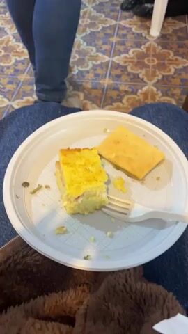 The camera pans from a plate of food, consisting of cornbread and a dish resembling a savory bread pudding, to a living room scene with eight people. The floor is tiled with a blue and yellow pattern. Several people are seated on sofas, while others are on chairs. A low wooden table sits in the center of the seating area. A large yellow painting of fish hangs on the wall. There's a surfboard leaning against the wall. One person is gesturing and speaking, while others are listening or talking amongst themselves. Many are holding drinks or food. The scene appears to be a casual gathering in a home.