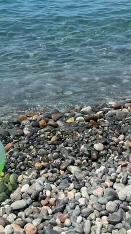 A close-up, eye-level shot shows a pebbly beach with the ocean waves lapping at the shore. A light green object, possibly a ball, is visible on the left side of the frame. In the foreground, a part of a circular, inflatable object with pastel colors, including pink and purple, and glittery accents is visible. Further out in the water, there are several people swimming or floating. The water is clear and blue, and the sky is bright and sunny.