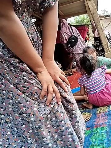 The video shows a group of people, including children and adults, gathered outdoors in what appears to be a rural setting. The scene is set under a wooden shelter with a corrugated metal roof. In the foreground, a person wearing a floral dress is partially visible. In the middle ground, two young girls are sitting on a colorful mat. One girl is wearing a striped top and a pink skirt, and the other is wearing a green t-shirt and dark shorts. Both girls appear to be interacting, with the girl in the pink skirt leaning forward. Several other people, including children and adults, are visible in the background, sitting and observing the scene. Some are dressed in traditional clothing. The ground is covered with mats and scattered objects, possibly shoes or other belongings. The overall atmosphere seems informal and communal.