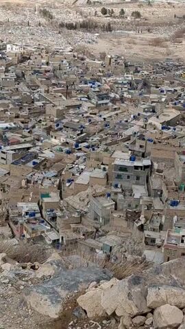 The video shows an aerial shot of a densely populated, somewhat impoverished urban area. Buildings are mostly low-rise, boxy, and constructed from light-colored materials. Many rooftops have blue water tanks. Beyond the urban area, a cemetery is visible, along with some sparse trees and a road. The foreground shows rocky terrain and sparse vegetation. No people are visible. The camera is stationary.