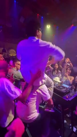 The video shows a crowded nightclub with a DJ booth. A person is being carried on the shoulders of another person in front of the DJ equipment. The crowd is dancing and raising their hands. The lighting changes from blue to red and purple. There are speakers and other DJ equipment visible on the booth. The overall atmosphere appears to be energetic and celebratory.