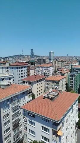 This video shows an aerial view of a dense urban cityscape on a clear day. The foreground features several multi-story residential buildings with terracotta tiled roofs and white facades. Beyond these, a variety of other buildings, some modern with glass exteriors and others older with muted colors, fill the landscape. A tall communication tower is visible in the distance, along with other skyscrapers and a vast expanse of urban development stretching to the horizon. The sky is a brilliant, cloudless blue. There are no people or animals visible in the frame, and no specific actions or interactions are taking place. The environment appears to be a typical residential and commercial urban area bathed in bright sunlight.