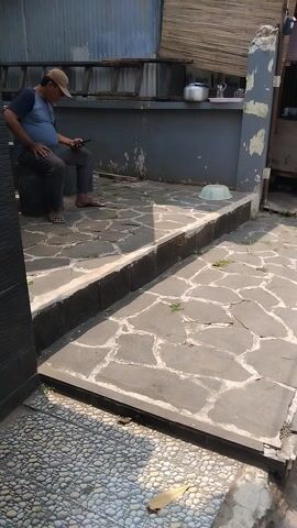 The video shows a man sitting on a step, looking at his phone. He is wearing a blue t-shirt, grey pants, and a tan baseball cap. He is barefoot. The scene is outdoors, with a stone-tiled patio in the foreground and a grey wall behind it. To the left of the wall, there is a wooden structure with bamboo blinds. Further back, there's a glimpse of a street with a motorcycle, a blue vehicle, and buildings decorated with colorful flags. The lighting suggests it is daytime, likely late afternoon or early morning given the long shadows. The overall impression is a casual, everyday scene in a residential or small commercial area, possibly in a tropical climate. There is no discernible text in the video.