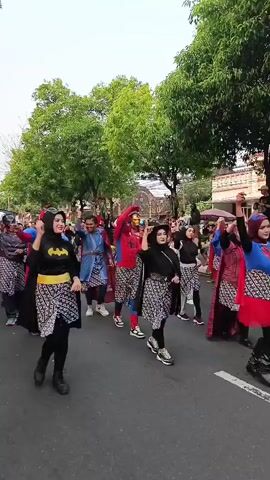 A group of people are walking in a parade, dressed in superhero costumes. The costumes include Batman, Superman, Spider-Man, Ultraman, and Black Panther. The people are also wearing traditional Indonesian patterned skirts. The parade is taking place on a street lined with trees and buildings. There are spectators watching the parade from the side of the street. The overall atmosphere appears to be festive and celebratory.
