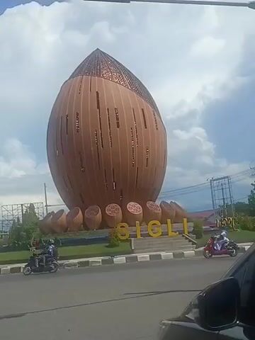 The media shows a large, brown, oval-shaped monument in Sigli, Indonesia, with a patterned, copper-colored top. The monument is surrounded by a circular garden with several smaller, also oval-shaped objects arranged at its base, and the word "SIGLI" is displayed in yellow lettering in front of the monument. Two people on a scooter are visible driving on the road in front of the monument. The sky is cloudy, suggesting it is daytime. The scene appears to be a public space, likely a roundabout or a park, given the presence of the road and traffic light. The monument's unique design, resembling a seed or nut, is a prominent feature of the landscape.