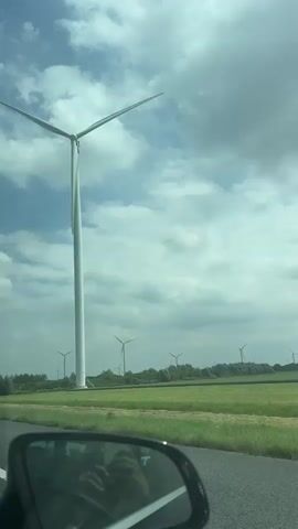 A car is driving down a road with a field of wind turbines in the background. The sky is cloudy. In the foreground, the side mirror of the car is visible, reflecting the person holding the camera. There are speed limit signs on the side of the road. One sign shows 100 km/h, and another shows 120 km/h from 6 AM to 7 PM. The video captures the view from inside the car as it passes by the wind turbines.