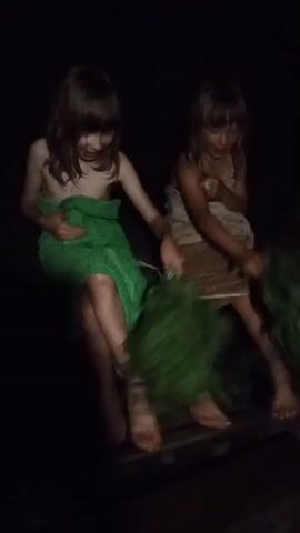 In a dimly lit, possibly outdoor setting that appears to be Buraly, Russia, two young girls are engaged in a ritualistic activity. Both are wearing towels around their waists, indicating a possible sauna or bathhouse experience. The girl on the left is draped in a bright green towel, while the girl on the right wears a patterned towel. They are both holding bunches of leafy branches, likely birch or oak branches, which are traditionally used in Russian banyas for massage and aromatherapy. The branches are being swung and potentially used to gently strike their bodies, a practice known as "venik" or "aromatherapy whisking."

The lighting is very low, suggesting it might be nighttime or a very dark interior. The focus is on the girls and their activity, with the background fading into blackness. The girls' faces are not clearly visible due to the low light and the motion of the branches, but their postures suggest a focused and perhaps playful engagement with the tradition. The presence of the veniks and the towels strongly points to a cultural practice associated with bathing and relaxation. There is no visible text in the media.