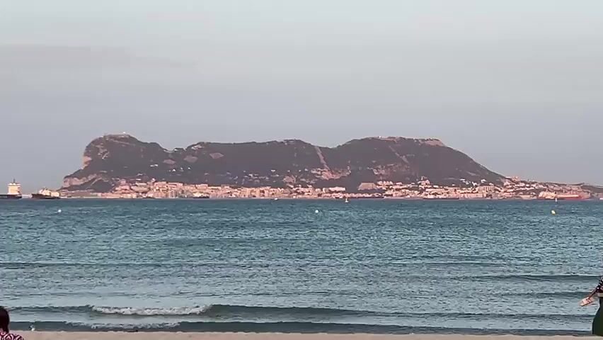 The video shows a wide shot of a beach scene with the sea in the foreground and a large rocky mass with a town built on its side in the background. Several people are visible on the beach, walking towards the right of the frame. The sea is blue with small waves rolling onto the sandy shore. The sky is a pale, clear color. There are ships visible in the distance on the water.