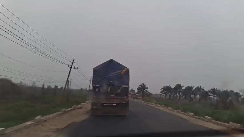 A truck travels down a wet, paved road under a cloudy sky. The truck is covered with a blue tarp. There are power lines on the left side of the road and palm trees on the right. A motorcycle with two people passes the truck. The road appears to be under construction, with piles of sand and gravel visible on the shoulder.