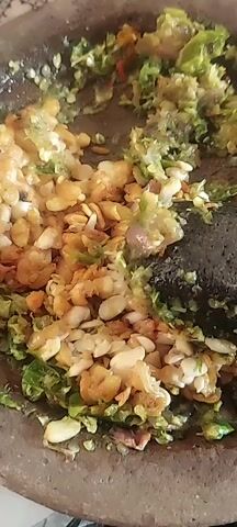 A close-up shot shows a mortar and pestle filled with ingredients. The mortar is made of dark-colored stone and contains a mixture of green chilies, what appear to be red chilies, pieces of onion, and small, light-colored beans or nuts. A dark stone pestle is positioned within the mortar, actively engaged in grinding the ingredients. The lighting suggests an indoor setting, possibly a kitchen. The camera angle provides a top-down perspective of the mortar and its contents.