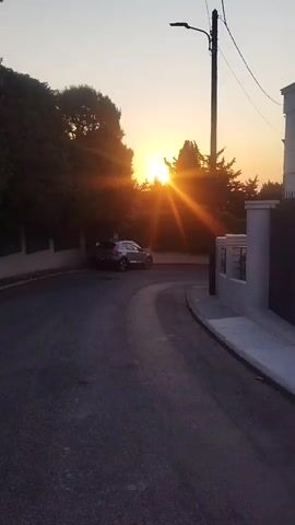 The video shows a street at sunset. The sun is visible in the distance, casting orange rays through the trees. A silver car is parked on the side of the road. A lamppost stands on the right side of the frame. The road is paved and curves to the right.