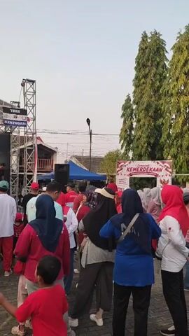 A group of people are gathered outdoors, facing a stage. The stage is set up with a large banner that reads "SEMARAK KEMERDEKAAN REPUBLIK INDONESIA". There is a speaker on the stage and a performer or presenter interacting with the audience. Many of the attendees are wearing red or white clothing, and some are wearing headscarves. The atmosphere appears to be festive, likely celebrating Indonesian Independence Day. The background shows buildings, trees, and a clear sky.