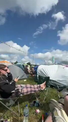 Here's a description of the video:

The video shows an outdoor scene at what appears to be a music festival or camping event. Several tents of various colors are pitched on a grassy field under a partly cloudy, blue sky. A string of colorful pennants hangs above the tents.

In the foreground, three people are relaxing in camping chairs. One person is wearing plaid pants and using a phone. Another person is wearing a striped shirt and also using a phone. Only the legs of the third person are visible. Several water bottles and other litter items are on the ground nearby.

The camera slowly pans upwards to show the tents and the sky. The overall impression is a relaxed, sunny atmosphere at an outdoor gathering.
