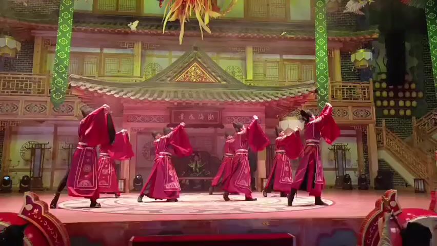 A group of eight performers dressed in red and black traditional Chinese costumes are dancing on a stage. They are positioned in a semi-circle in the center of the stage. The stage is decorated with an elaborate backdrop of a traditional Chinese palace or temple building. The performance appears to be a traditional Chinese dance. The lighting is theatrical and colorful. There are no other people visible besides the performers.