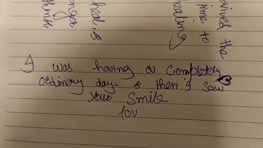 A hand holding a blue pen is writing on lined paper. The writing says "I was having a completely ordinary day - then I saw your smile love you." There are also some handwritten words in purple ink above the main message.
