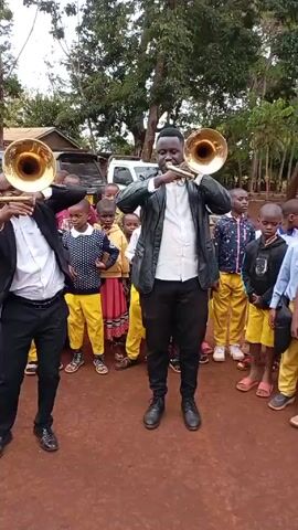 Here is a description of the video:

The video shows two men playing instruments in front of a group of young children.  The men are both dressed in jackets, white shirts and dark pants. The man on the left has a blazer while the man on the right is wearing a leather jacket.  Each of them is holding a brass instrument, and they appear to be playing a song for the children. The children are standing in a semi-circle around the men. They are dressed in a variety of clothes, including yellow pants.  

The background of the video shows trees and a building. The ground is red dirt.  The overall scene appears to be taking place outdoors.