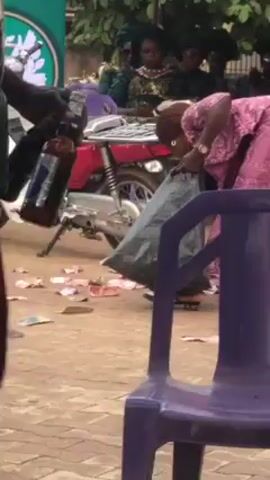 The video appears to show an outdoor event. Two men are featured prominently: one wearing a white shirt with blue floral patterns and red shorts, and another in a striped tank top. The man in the floral shirt seems to be handling bottles, possibly drinks. They appear to be near a purple plastic chair and some sound equipment. In the background, there are other people, some seated, under what appears to be a tent or canopy. A sign saying "Diamond Ladies Women Club" is visible. The ground seems to be an outdoor surface, possibly pavement or dirt.