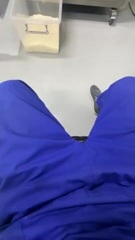 The video appears to be filmed from a first-person perspective, showing a person's legs clad in blue scrubs. The person is seated in a chair, and the camera pans across a clean, modern laboratory or cleanroom environment.

The scene features a sterile, white floor, white walls, and a white door with a window. There is a large window looking into another room, where some equipment and shelves are visible. In the foreground, on a counter, is a clear plastic container filled with what appears to be a light-colored granular substance, possibly food ingredients or lab materials.

The lighting is bright and even, suggesting an indoor setting during daytime. There are no obvious cues about the specific time of day beyond it being well-lit. The overall impression of the environment is one of cleanliness, order, and a focus on scientific or industrial processes. The person's attire suggests they are likely a worker or technician in this facility. No specific emotions are conveyed, but the movement of the camera suggests a tour, a transition between areas, or a moment of observation. There is no visible text within the video frames. The location context provided is Nalazar, Uzbekistan, which aligns with the clean and potentially industrial or research-oriented setting.