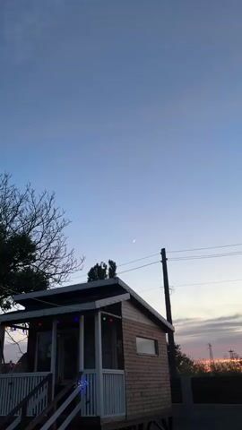 The video shows a crescent moon and a bright star in a pale blue twilight sky. Below, a small wooden shed with a sloped roof is visible, adorned with colorful lights. A dark tree branch extends into the frame from the left, and power lines stretch across the sky. The overall scene is tranquil and suggestive of early evening.