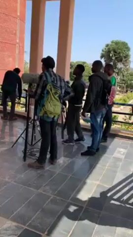 The scene shows a group of five men on a tiled patio with columns. Four of the men are standing, with one holding a sound boom and wearing a backpack. One man is bending over in the corner. One of the men walks away from the group to the left. The background includes a brick building, a few plants, and a low fence.