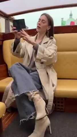 A woman is sitting on a yellow bench inside a boat with a transparent roof. She is wearing a tan trench coat, a grey sweater, blue jeans with a frayed hem, and beige heeled boots. She is holding a small black video camera up and recording herself. She has brown hair and hoop earrings. The boat appears to be moving along a canal.