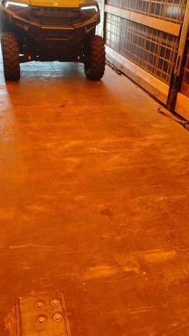 This media file features a close-up, low-angle, interior shot of a yellow off-road vehicle, possibly a UTV or side-by-side, parked on a textured, orange-toned surface. The vehicle's rugged tires and bright LED headlights are prominently displayed. The background is a dimly lit, industrial-looking space with a metal grate structure visible in the upper left corner, suggesting a garage, workshop, or possibly a vehicle transport.

The media file captures the vehicle from a perspective that emphasizes its size and robust design. The lighting is warm and artificial, casting distinct shadows. There is no visible text within the frame of the media file itself. The scene appears to be static, with the vehicle stationary. The overall impression is one of industrial utilitarianism, highlighting the vehicle's intended use for off-road adventures or heavy-duty tasks. There are no people visible, and the emotional tone is neutral, focusing on the object itself and its environment. Given the Valga, Estonia location, it is possible this is a commercial or personal vehicle within a facility in that area.