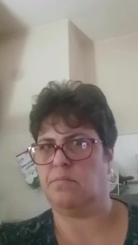 This is a close-up, slightly blurry video of a woman with dark, short hair and glasses. She is looking directly at the camera with a neutral expression. The background appears to be an indoor setting, possibly a kitchen or living area, with a light-colored wall and some indistinct objects. The video is very short, only a few seconds long.