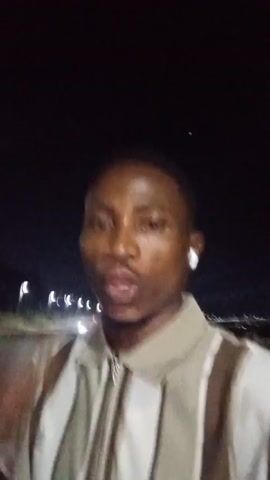 The video shows a man standing outside at night. He wears a brown and white striped zip-up shirt, a gold watch, and a gold ring. He gestures with his hands, touching his forehead, covering his mouth, and making a phone call gesture to the camera. A street light is visible in the background.