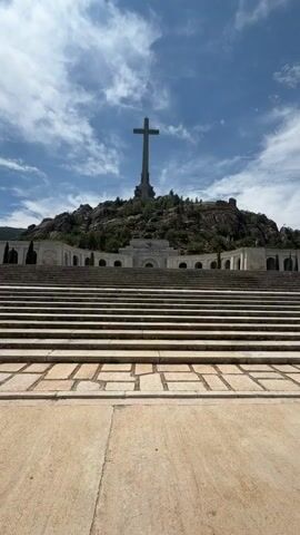 A low-angle, vertical, full shot video captures a monumental cross atop a rocky, tree-covered hill. In the foreground, a wide, stone staircase leads upwards. The sky above is a vibrant blue, dotted with fluffy white clouds. The scene appears to be outdoors, possibly a memorial site, under bright daylight conditions. No people or animals are visible in the frame. The overall impression is one of grandeur and solemnity.
