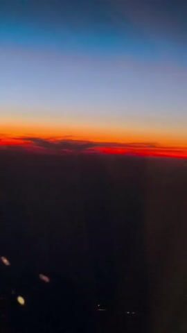 The video shows a view from an airplane window at sunset. The sky transitions from a deep blue at the top to orange and red near the horizon. Below the horizon, the landscape is dark, with faint lights visible on the ground. There are some clouds at the horizon that are illuminated by the setting sun. There are no people or distinct objects visible in the frame, other than what appears to be the window frame or reflections on the glass in the lower left corner.