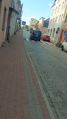 The video shows a street scene with buildings on both sides. A cobblestone sidewalk runs along the left side of the street, and a paved road with cobblestone sections is on the right. A pedestrian crossing is visible on the road. Several vehicles are present: a dark van, a red sports car, and another car further down the road. A person is walking on the sidewalk. The sky is blue with some clouds. A blue pedestrian crossing sign is posted on the left side of the street. The buildings appear to be of historical architecture, with awnings and windows visible. There are also some signs and advertisements on the buildings.