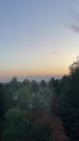 The video captures a serene sunset scene over a landscape dominated by lush green trees. The sky displays soft gradients of light blue, yellow, and subtle pink hues, indicative of dusk. In the distance, an airplane is visible, initially as a small silhouette, then becoming clearer as it approaches. Its landing lights are prominently illuminated, suggesting it is on a descent, possibly preparing to land. The plane moves from the center of the frame towards the left as it gets closer, its bright lights piercing the fading light of the sky. The foreground and midground are filled with the canopy of various trees, creating a dark, natural outline against the colorful sky. The overall impression is one of a peaceful evening with a visible sign of air travel. The location context provided is Otopeni, Romania.