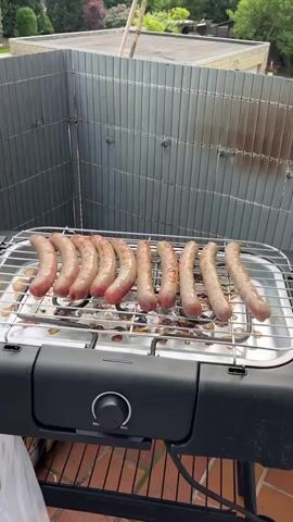 The video presents a barbecue scene on what appears to be a balcony or patio. Initially, a row of sausages is seen cooking on an electric grill. The camera then pans to a section of gray fencing. Next, the scene transitions to a cutting board with sliced beef, a bottle of Perlenbacher Pils beer, salt, a covered pot, a knife, and a fork. Finally, a wine glass filled with a yellow liquid is added to the scene, sitting alongside the beer. The setting suggests a relaxed outdoor dining experience.