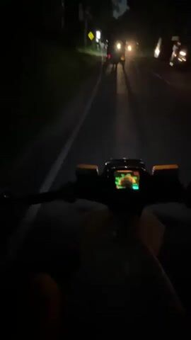 The video shows a first-person perspective of someone riding a motorcycle at night. The road is asphalt with a solid white line dividing the lanes. There are cars ahead with headlights on. A yellow sign is visible on the left side of the road. The motorcycle has a digital display and orange indicator lights. It is dark, and the view is illuminated by the motorcycle's headlight and the headlights of the vehicles in front.