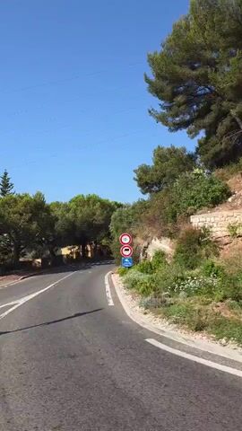 A roadside view with a clear blue sky. A paved road curves to the right. A traffic sign indicating directions to "A 8 (NICE)" and "TURBIE" is visible on the left, along with a smaller sign that appears to be a bus stop timetable. Green trees and foliage line the roadside, with a rocky embankment on the right. A few buildings are visible in the background. The sun is shining, casting shadows on the road.