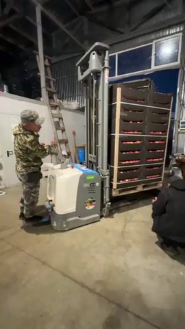 A man in camouflage clothing operates a powered pallet jack inside a warehouse. He is facing a stack of dark brown crates filled with apples, which are loaded onto a pallet. The pallet jack is lifting the pallet of apples. Another man in a black jacket and hood is crouching near a white wall to the right of the scene. A ladder leans against a corrugated metal wall on the left side of the frame. The warehouse appears to be a cold storage facility, suggested by the white insulated wall on the right and the metallic appearance of the structure. The lighting is dim, with artificial lights visible overhead. The overall environment suggests a work setting related to food storage or distribution.