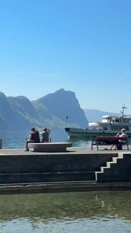 This video presents a picturesque daytime scene in Vitznau, Switzerland, showcasing a serene lake and mountain landscape under a bright, clear blue sky.

In the foreground, a concrete pier or dock structure extends into the calm, clear water, revealing the lakebed beneath. On the left side of this pier, two individuals are seated on a low concrete bench, facing out towards the lake and the distant mountains. Further along the pier to the right, another person is seen relaxing on a red park bench.

A prominent feature in the mid-ground is a large white passenger boat or ferry, which is actively moving across the lake. The boat has multiple decks and appears to be carrying passengers enjoying the scenic journey. A small flag, identifiable as the Swiss flag, is visible on the boat's mast, confirming its location.

The background is dominated by majestic, verdant mountains, with some rocky outcrops visible, creating a dramatic and beautiful backdrop to the expansive lake. The water in the main body of the lake is a deep blue, contrasting with the clearer, shallower water closer to the pier.

The overall atmosphere conveyed is one of tranquility and natural beauty, suggesting a pleasant and relaxing day by the lake.